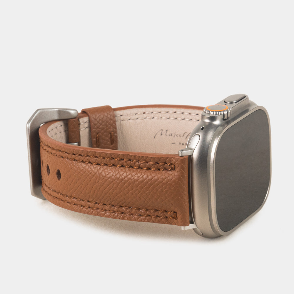 Apple Watch Ultra bandje heren 49 mm goud Epsom leer met titanium gespen — driekwart profiel op de titanium Ultra-kast, crèmekleurige Alsavel voering met stempel Marcel Robert Paris