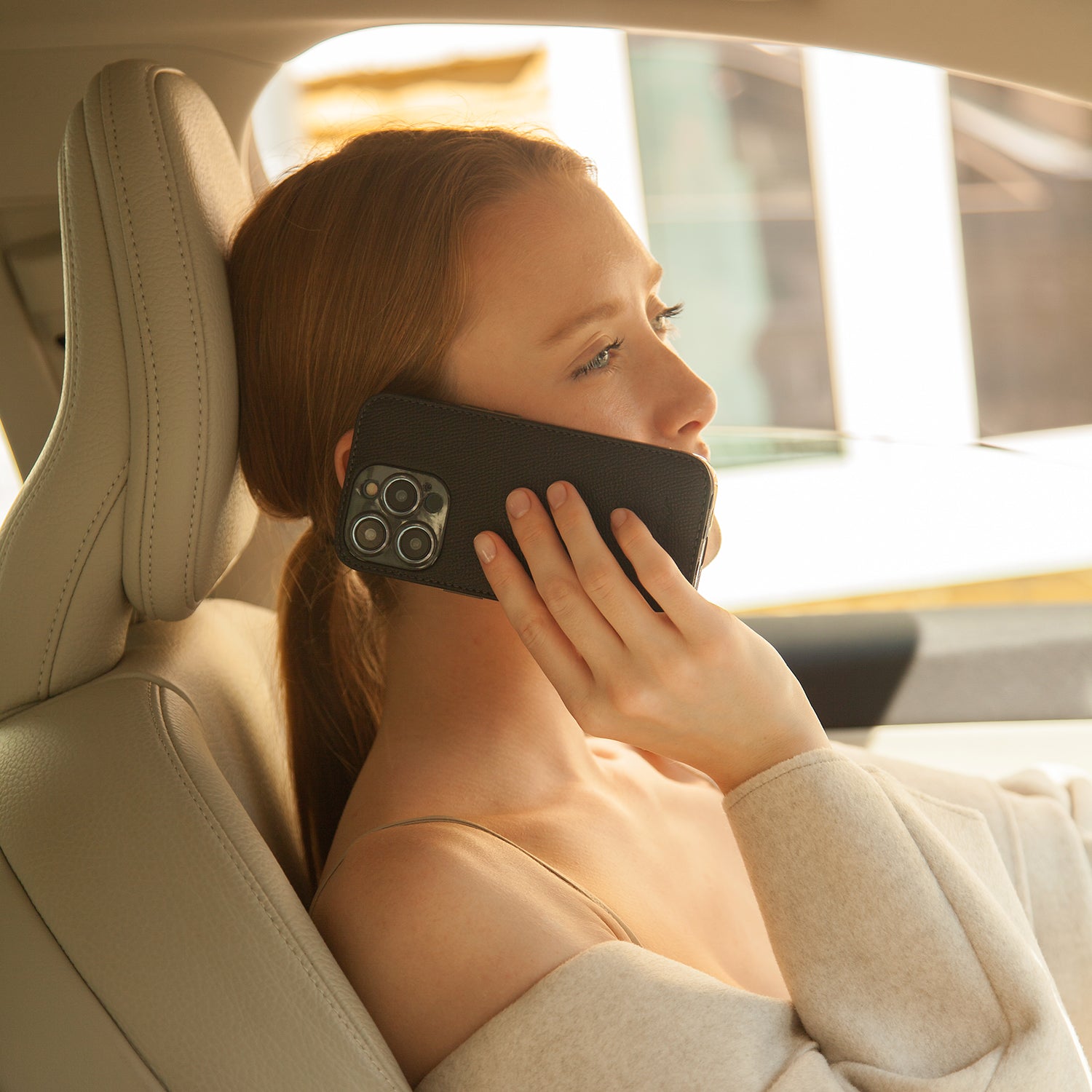 Woman sitting in a car, using a phone in black leather case with a sunset or sunrise in the background