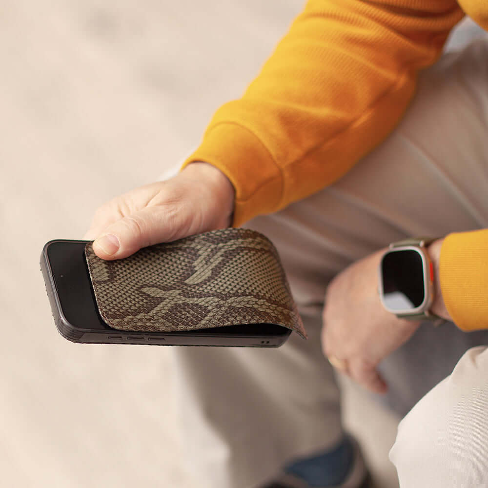 Person holding a phone with a case made of python leather, wearing a yellow sleeve and white pants.