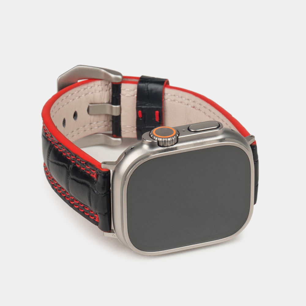 Apple Watch Ultra 49 mm aus schwarzem Alligatorleder, Naht und Kante in Rot, mit Adaptern und Schnalle aus Titan | 3/4-Frontansicht mit Titangehäuse und den quadratischen Schuppen des Leders