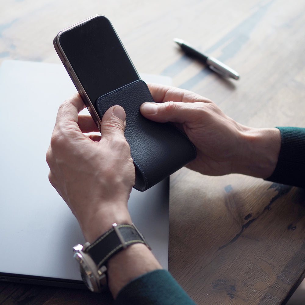 Blue calfskin leather flip case for iPhone 15 held in both hands over a desk, flap folded open behind the phone