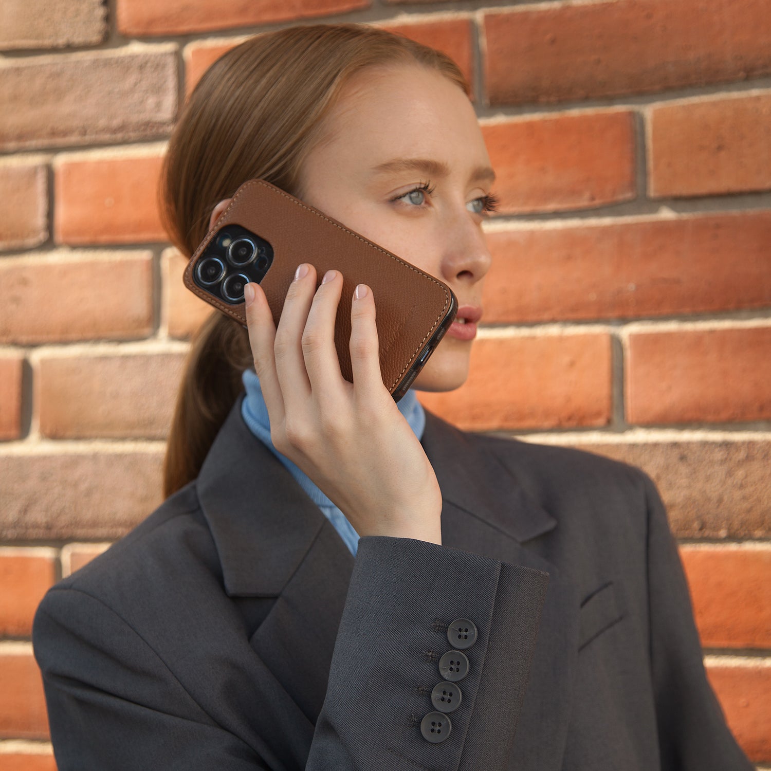Woman taking a call against brick wall with iPhone 15 Pro Max in Marcel Robert gold Saffiano calfskin leather cover case
