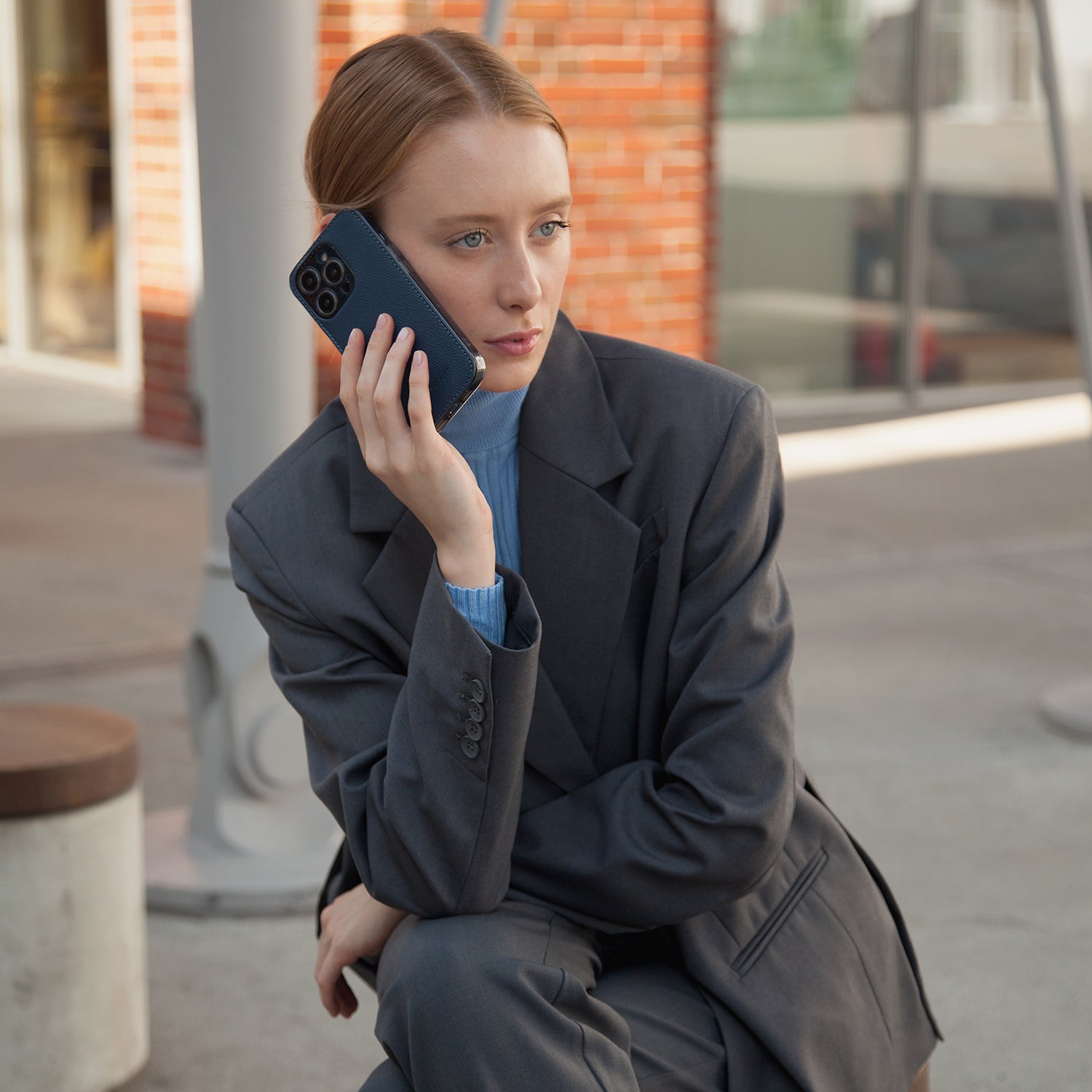 Woman in a dark coat sitting outdoors, holding a phone in leather cover case to her ear.