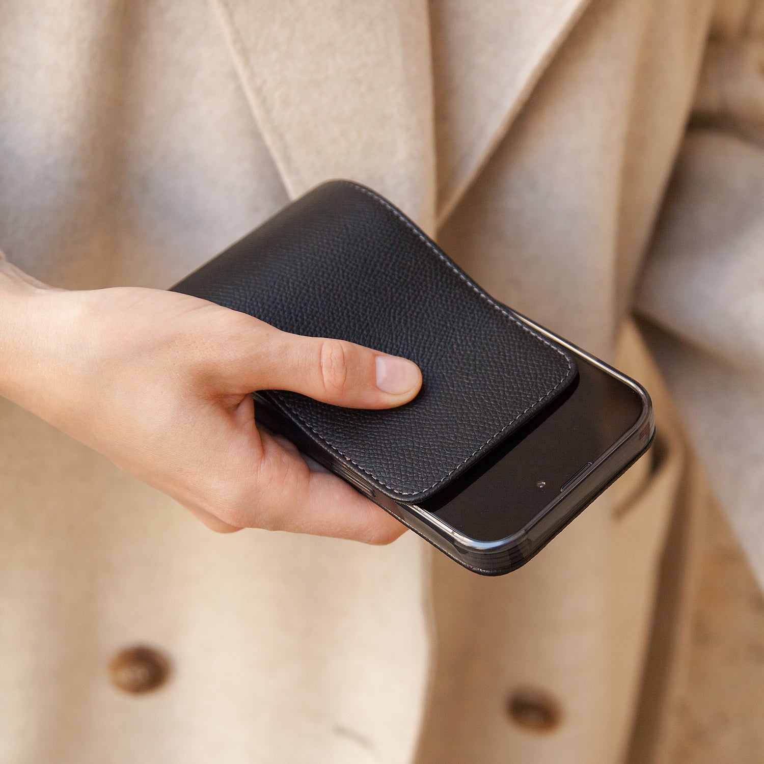 Hand holding a phone in black leathrt case against a beige coat background