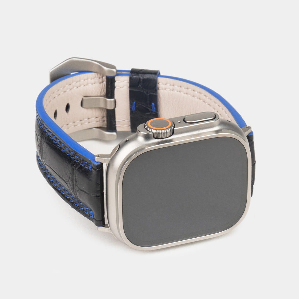 Apple Watch Ultra 49 mm aus schwarzem Alligatorleder, Naht und Kante in Blau, mit Adaptern und Schnalle aus Titan | 3/4-Frontansicht mit Titangehäuse und den quadratischen Schuppen des Leders