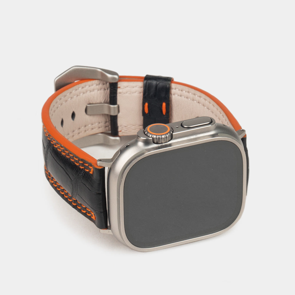 Apple Watch Ultra 49 mm aus schwarzem Alligatorleder, Naht und Kante in Orange, mit Adaptern und Schnalle aus Titan | 3/4-Frontansicht mit Titangehäuse und den quadratischen Schuppen des Leders