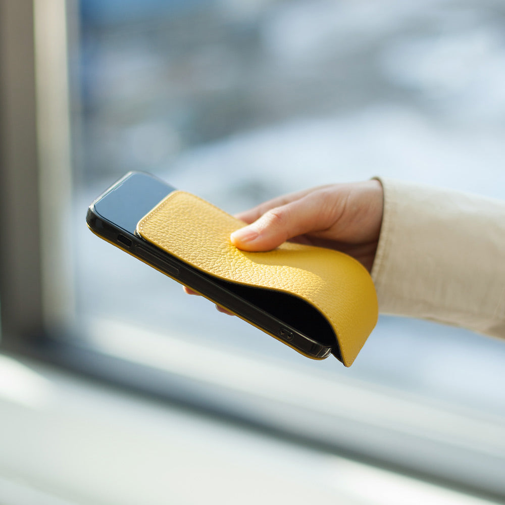 Cover in pelle di vitello gialla per iPhone 15 Plus tenuta in una mano davanti a una finestra, patta ripiegata dietro il telefono