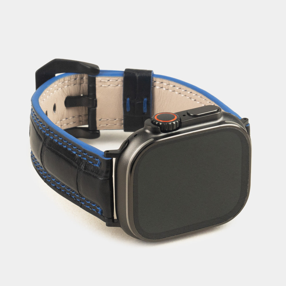 Apple Watch Ultra 49 mm aus schwarzem Alligatorleder, Naht und Kante in Blau, mit schwarzen Adaptern und Schnalle | 3/4-Frontansicht mit schwarzem Gehäuse und den quadratischen Schuppen des Leders