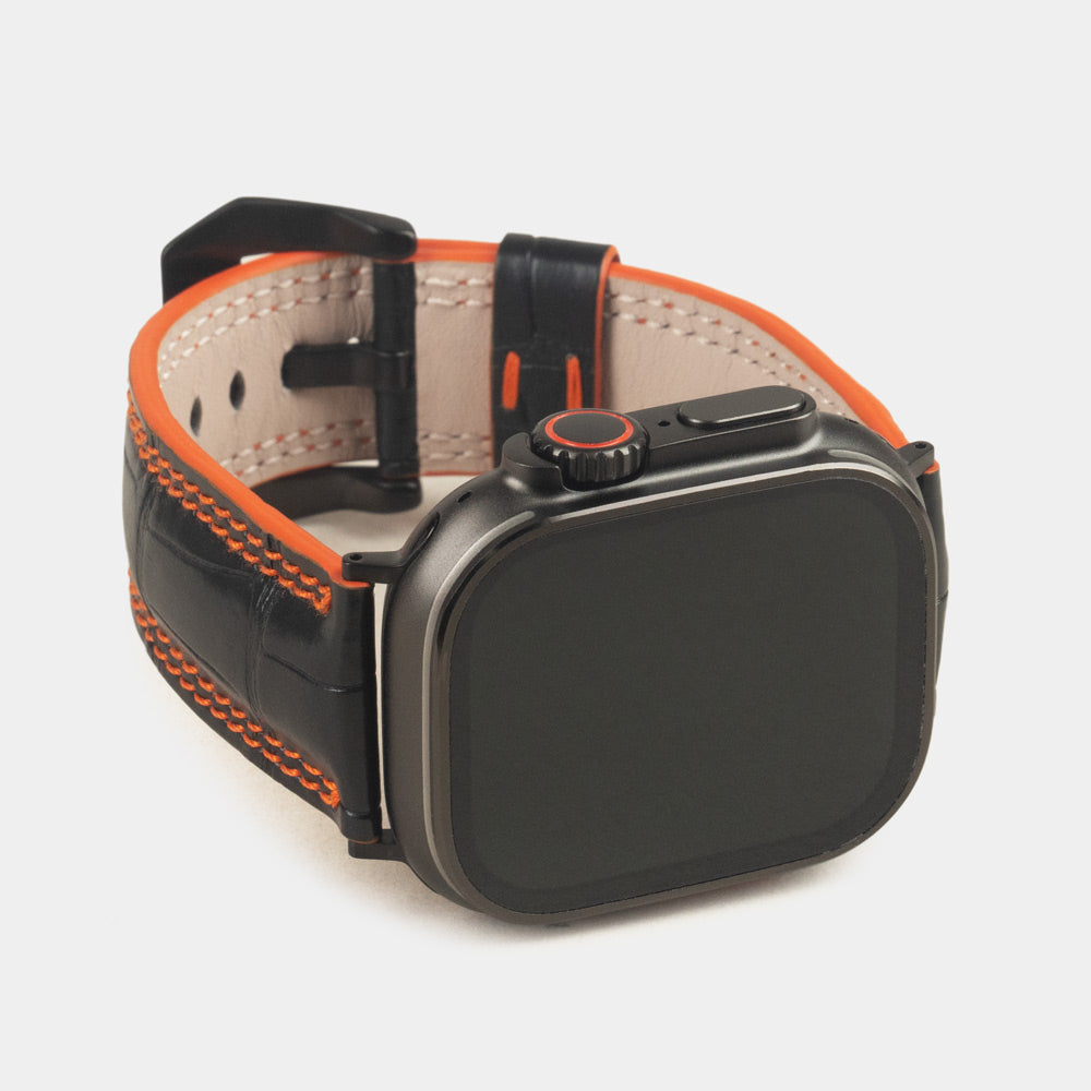 Apple Watch Ultra 49 mm aus schwarzem Alligatorleder, Naht und Kante in Orange, mit schwarzen Adaptern und Schnalle | 3/4-Frontansicht mit schwarzem Gehäuse und den quadratischen Schuppen des Leders